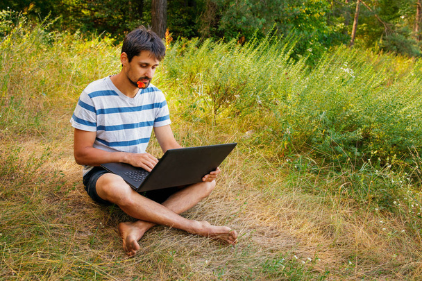 A young man sits on the grass with a flower in his mouth and works correspondence on a laptop. - Foto, Bild