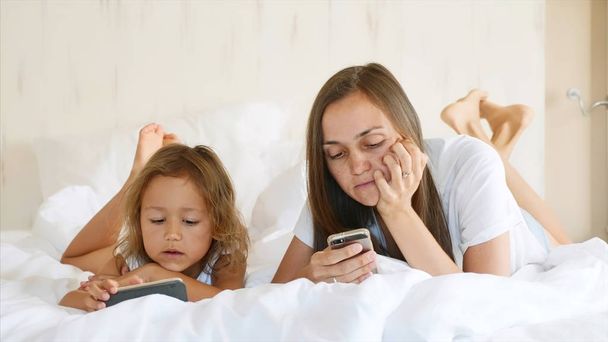 Young mother with little cute daughter are using gadgets lying on a white bed. - Fotó, kép