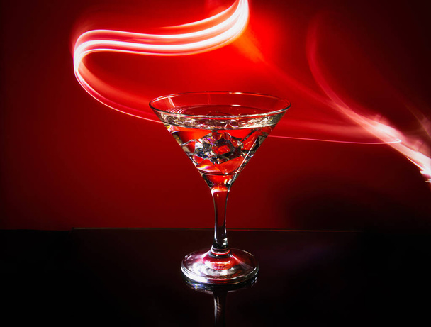 Sklenice s Martini izolovaná na červeném pozadí s proužky neonové světelné malby za nimi. Světelná vlna, přehrávání barev, kreslení světlem. - Fotografie, Obrázek