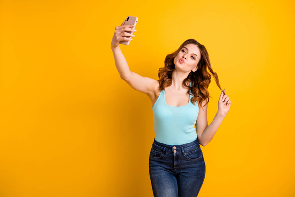 Foto van Lady houden telefoon maken Self Foto's met Coquette Kisses slijtage Tank-Top en jeans geïsoleerd gele achtergrond - Foto, afbeelding