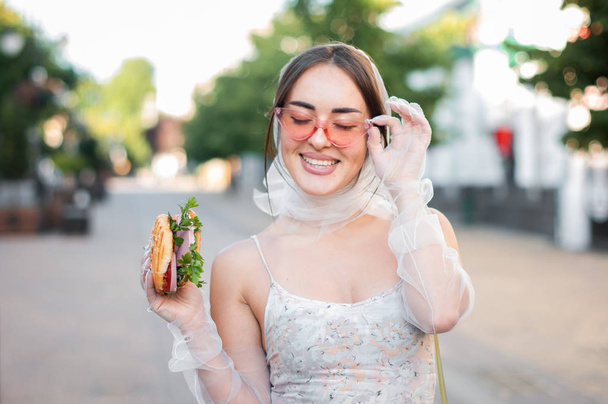Charming girl in pink sunglasses and a dress eats a hamburger on the street - Photo, Image
