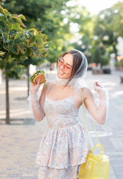Charming girl in pink sunglasses and a dress eats a hamburger on the street - Photo, Image