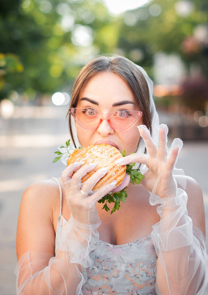 Charming girl in pink sunglasses and a dress eats a hamburger on the street - Foto, afbeelding