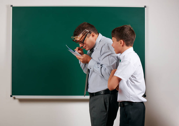 Portrait of a teacher checking homework, reading school exercise books and schoolboy posing on blackboard background - back to school and education concept - Photo, Image