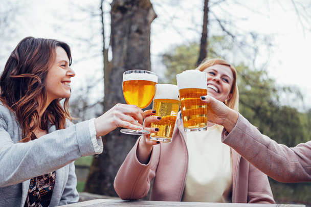 kolme naista istuu syksyllä ulkoterassilla puhumassa, nauramassa ja paahtamassa yhdessä oluen kanssa
. - Valokuva, kuva