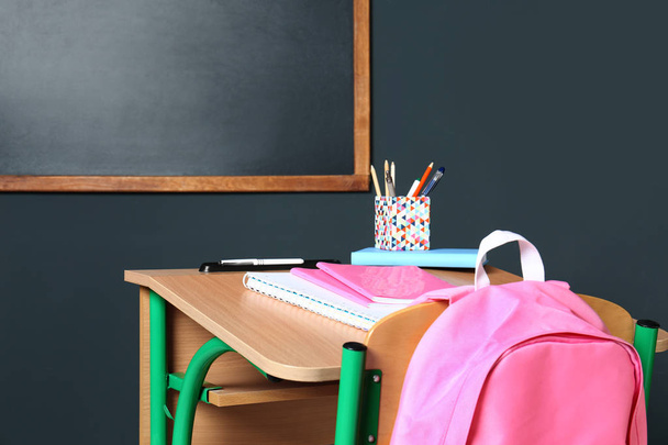 Дерев'яний шкільний стіл з канцтоварами та рюкзаком біля дошки на сірій стіні
 - Фото, зображення