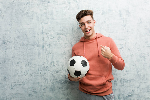 Молодой спортсмен, держа футбольный мяч, удивился, указывая на себя, широко улыбаясь
. - Фото, изображение