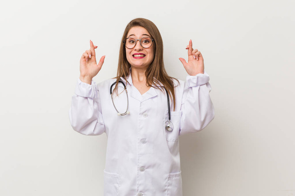 Jonge dokter vrouw tegen een witte muur kruising vingers voor het hebben van geluk - Foto, afbeelding