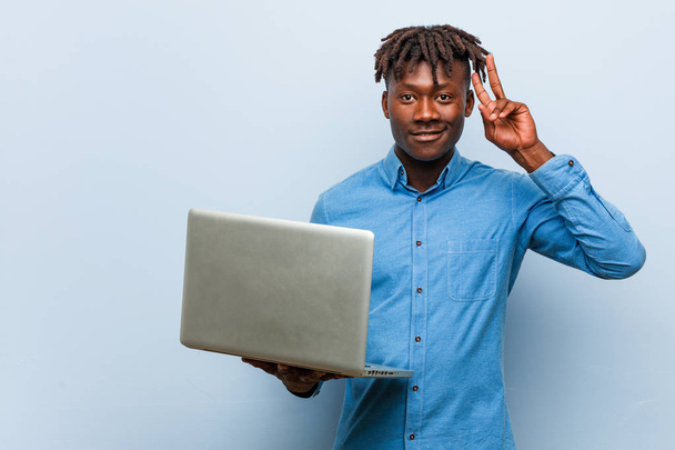Young rasta black man holding a laptop showing victory sign and smiling broadly. - 写真・画像