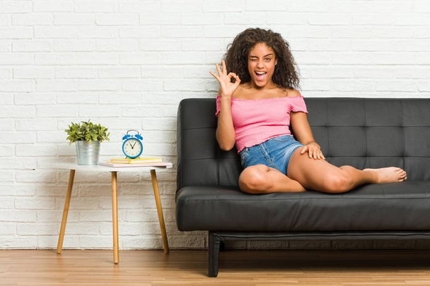 Joven mujer afroamericana sentada en el sofá guiña un ojo y sostiene un gesto bien con la mano
. - Foto, imagen