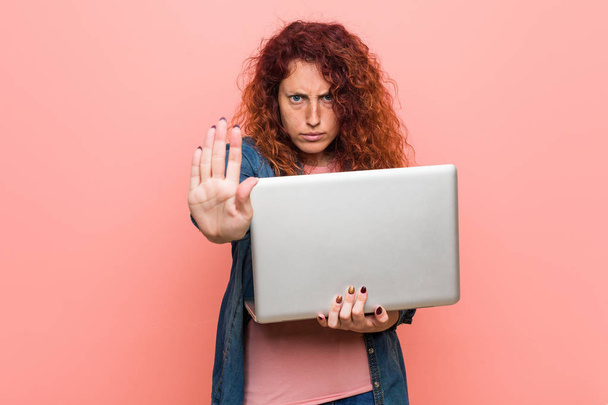 Nuori valkoihoinen punapää nainen, jolla on kannettava tietokone, jossa on ojennettu käsi, joka näyttää stop-merkkiä, estäen sinua
. - Valokuva, kuva