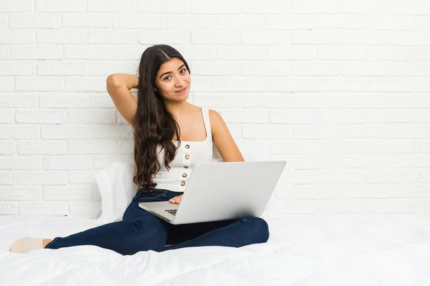 Νέα αραβική γυναίκα που συνεργάζεται με το laptop της στο κρεβάτι αγγίζοντας πίσω από το κεφάλι, σκεπτόμενος και κάνοντας μια επιλογή. - Φωτογραφία, εικόνα