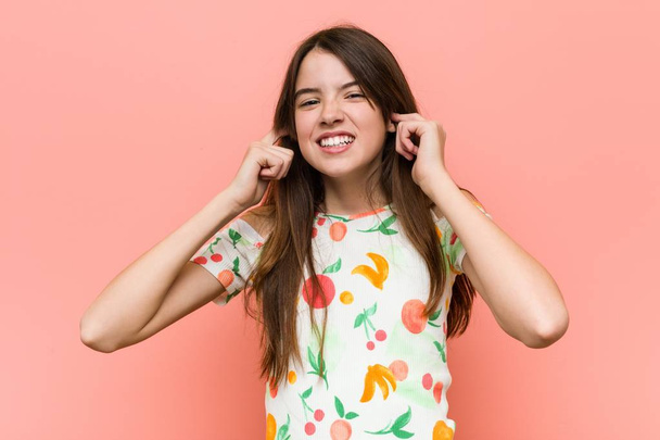 Chica usando una ropa de verano contra una pared roja cubriendo las orejas con las manos
. - Foto, imagen