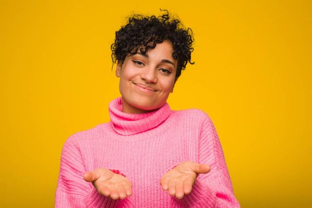 Giovane donna afroamericana che indossa un maglione rosa che tiene qualcosa con le palme, offrendo alla fotocamera
. - Foto, immagini