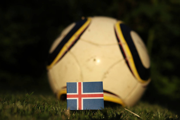 Κρατική σημαία της Ισλανδίας σε ξύλινο ραβδί μαχαιρωμένο στην επιφάνεια του χόρτου. Στο παρασκήνιο υπάρχει χαρακτηριστική μπάλα για το παιχνίδι. Παγκόσμιο Πρωτάθλημα 2022. Ευρώ 2020 - Φωτογραφία, εικόνα