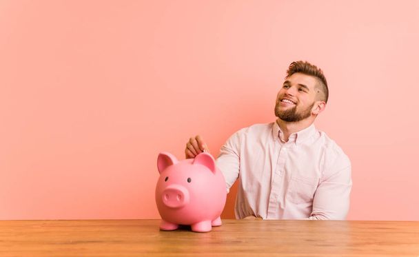 Young caucasian man sitting with a piggy bank smiling confident with crossed arms. - 写真・画像