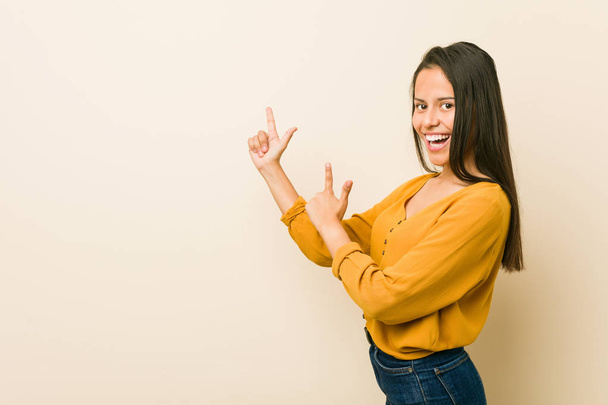 Young hispanic woman against a beige background pointing with forefingers to a copy space, expressing excitement and desire. - Foto, immagini