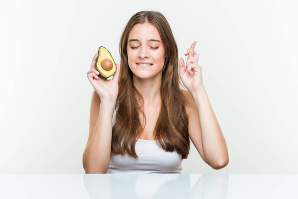 Young caucasian woman holding an avocado crossing fingers for having luck - Photo, Image