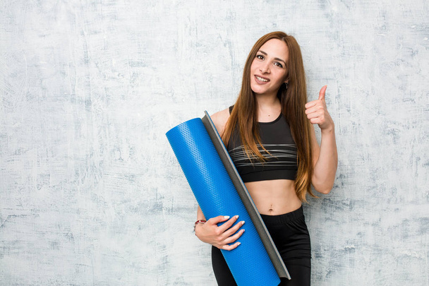 Young caucasian sportswoman holding a mat smiling and raising thumb up - Photo, Image