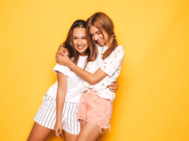 Two young beautiful smiling hipster girls in trendy summer clothes. Sexy carefree women posing near yellow wall in studio. Positive models going crazy and having fun.Hugging - Photo, Image