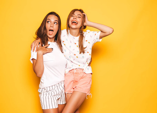 Two young beautiful smiling hipster girls in trendy summer clothes. Sexy carefree women posing near yellow wall in studio. Positive models going crazy and having fun.Hugging - Foto, afbeelding