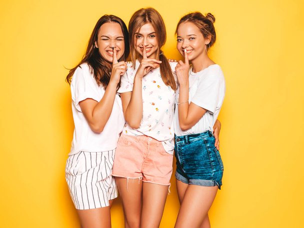 Three young beautiful smiling hipster girls in trendy summer clothes. Sexy carefree women posing near yellow wall in studio. Positive models showing hush finger silence sign, gesture - Foto, imagen