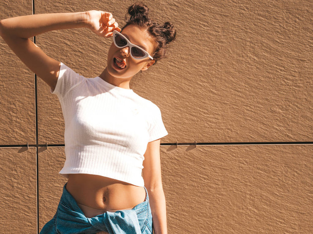 Beautiful smiling model with horns hairstyle dressed in summer hipster white t-shirt clothes.Sexy carefree girl posing in the street near wall.Trendy funny and positive woman having fun in sunglasses - Photo, image