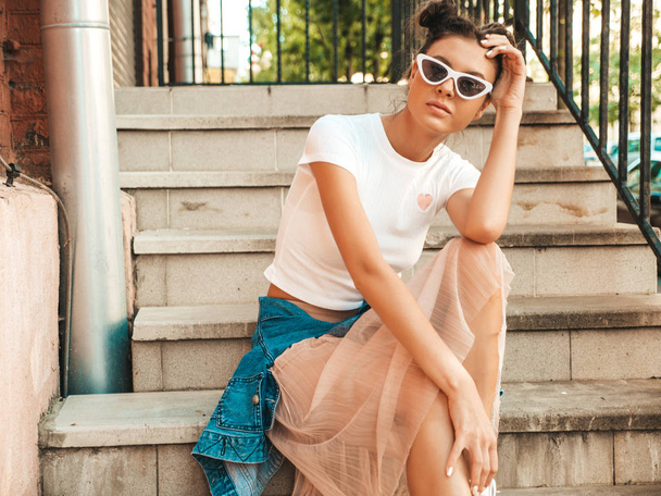 Beautiful model with horns hairstyle dressed in summer hipster jacket jeans clothes.Sexy carefree girl sitting on stairs in the street.Trendy funny and positive woman having fun in sunglasses - Photo, image