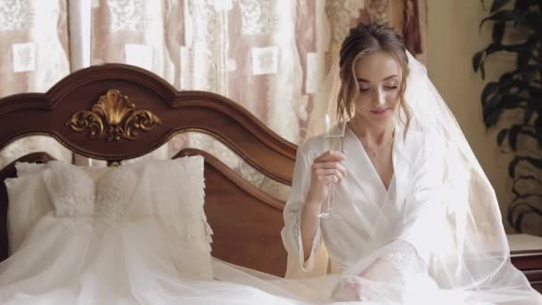 Mooie en mooie bruid in nachtjapon en sluier zitten en drinken champagne - Video