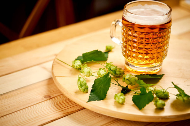 Large pint of beer with foam and sprigs of hops on wooden table - Фото, изображение