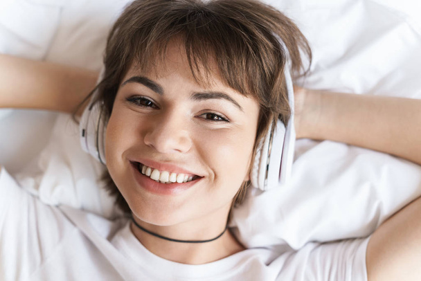 Optimistic positive pleased young beautiful woman lies indoors at home on bed listening music with headphones. - Foto, Bild