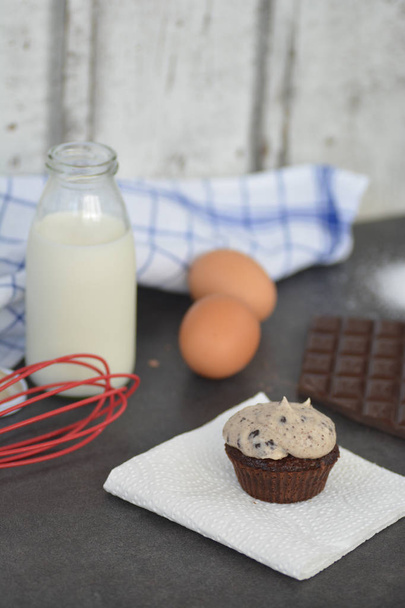 Homemade muffin with cream topping and chocolate, milk, eggs and flour on a kitchen surface - Photo, image