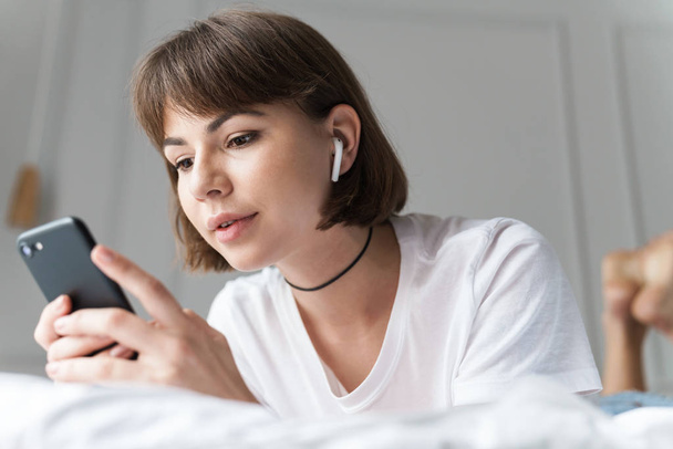 Concentrated young pretty woman indoors at home listening music with bluetooth earphones using mobile phone. - Photo, Image