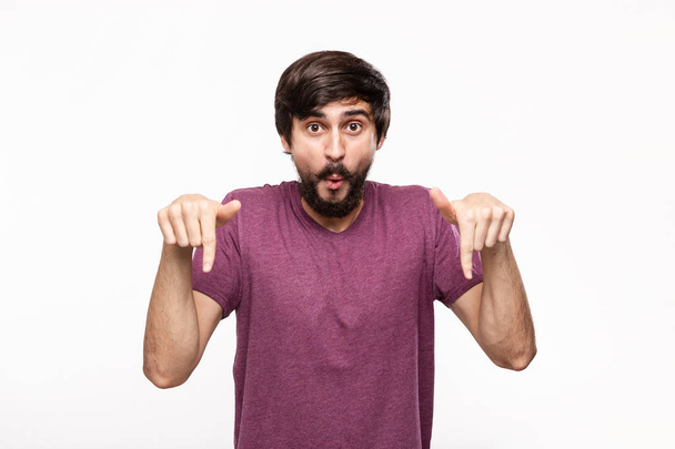 Excited brunet man in a purple shirt with beard and mustaches pointing with his fingers down standing isolated over white background. Place of product and advertisment. - Photo, Image