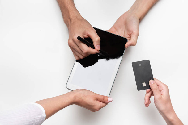 Close up top view of a woman hands over bank card to a man in order to pay for the product. Tablet and pen on a white table. Concept of e-commerce and online shopping. Successful deal. - Foto, Bild
