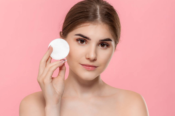 美しいモデルは、白い綿のスポンジで肌に化粧品を適用します。モデルは、ピンクの背景の上に分離されたカメラを見ます。美容・健康治療の概念. - 写真・画像