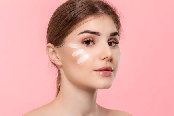 Beautiful model applyes moisturizing cosmetic product to skin holding white tube of a hyaluronic cream isolated over pink background. Concept of beauty and health treatment. - Фото, изображение