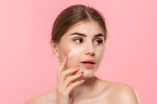 Beautiful model applyes moisturizing cosmetic product to skin holding white tube of a hyaluronic cream isolated over pink background. Concept of beauty and health treatment. - Valokuva, kuva