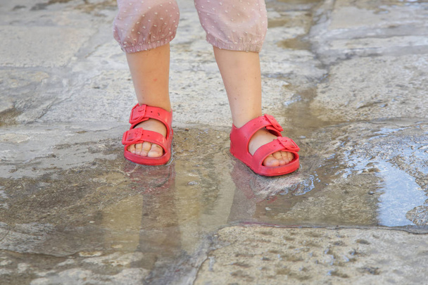 Детские ножки в красных резиновых сандалиях прыгают в лужу дождя
 - Фото, изображение