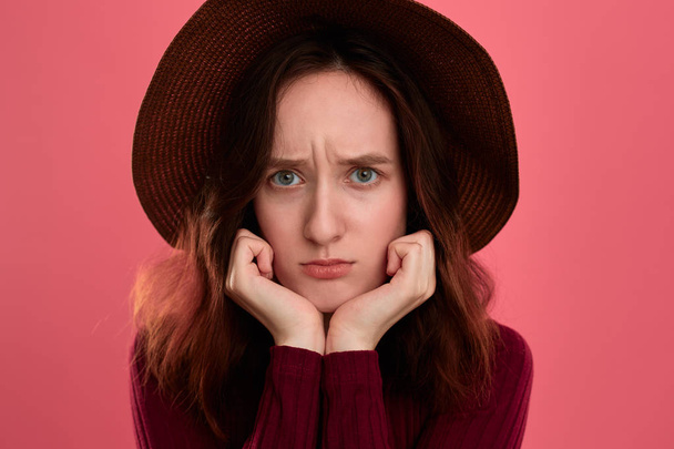 Close-up portrait of an upset beautiful brunette girl in a wide-brimmed hat posing on a dark pink background. - Photo, Image