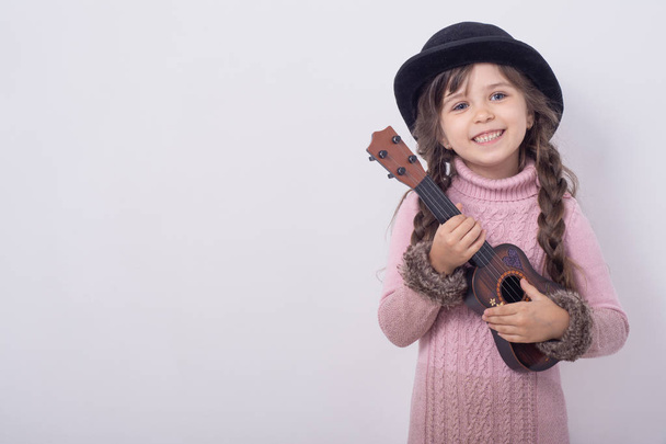 Ένα μικρό κορίτσι που έπαιζε κιθάρα σε ένα γκρίζο φόντο τοίχου. Αστείο κοριτσάκι με κιθάρα από το γιουκαλέλι. Μοντέρνο κορίτσι κάντρι παίζει μουσική.  - Φωτογραφία, εικόνα