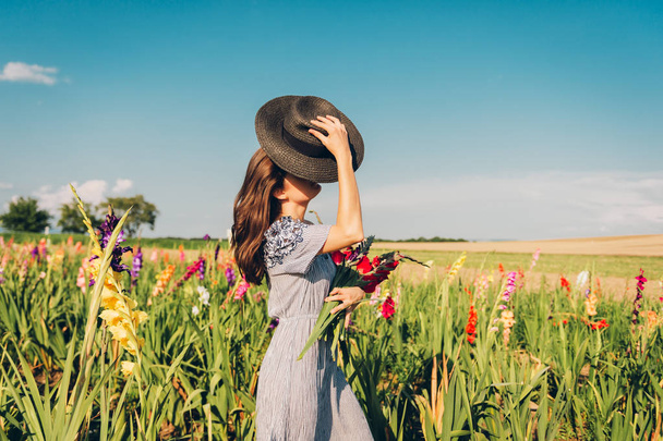 Gorgeous young woman picking flowers in a field, wearing summer dress, black straw hat  - Fotoğraf, Görsel