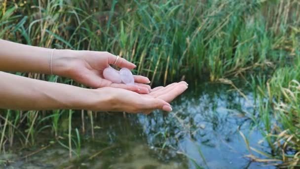Female hand holding a rose quartz and amethyst crystal yoni eggs on river background. Womens health, unity with nature concepts - Footage, Video