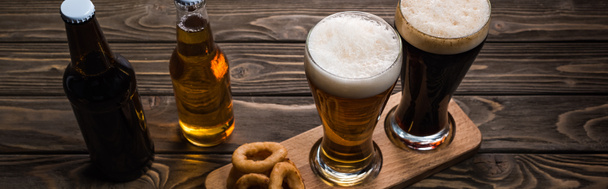 panoramic shot of glasses and bottles of dark and light beer near fried onion rings on wooden table - Fotoğraf, Görsel