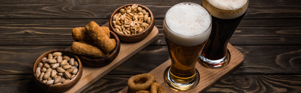 панорамный снимок стаканов тёмного и светлого пива возле чаш с арахисом, фисташками, жареным сыром и луковыми кольцами на деревянном столе
 - Фото, изображение