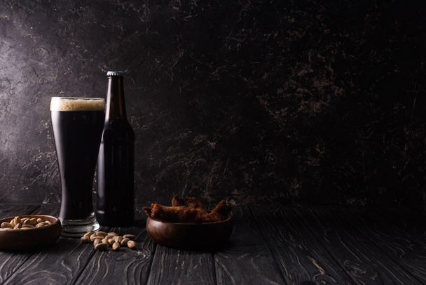vaso y botella de cerveza oscura cerca de cuencos con pistachos y alas de pollo sobre mesa de madera
 - Foto, Imagen
