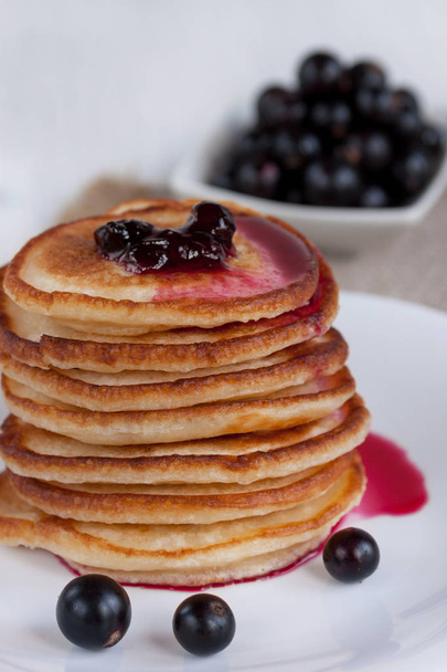 Stack of american pancakes with blackcurrant jam and fresh blackcurrant berries are lying on a white plate. - Фото, зображення