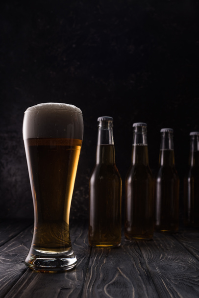 glass of fresh light beer with foam near bottles on wooden table - Foto, afbeelding