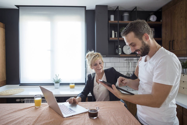 Couple in kitchen in the morning. They are smiling and browsing tablet and laptop. - Photo, Image