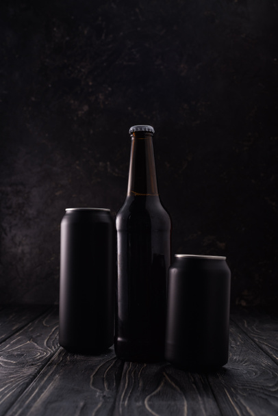 φιάλη μεταξύ μαύρων μεταλλικών κουτιών μπύρας σε ξύλινο τραπέζι  - Φωτογραφία, εικόνα